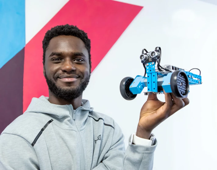 Student in a grey hoodie holding up a blue robotic vehicle, smiling against a colourful geometric wall. Student in a grey hoodie holding up a blue robotic vehicle, smiling against a colourful geometric wall.